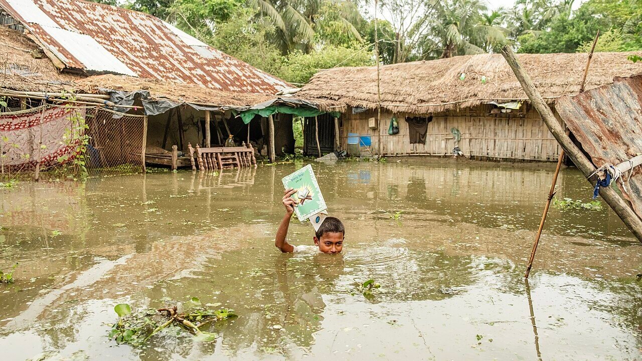 A child holding a book above floodwater outside submerged homes